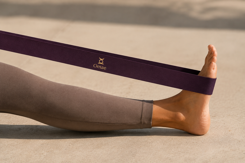 A close-up of a person's foot and lower leg resting on a beige floor, wearing a dark purple yoga strap with a gold logo that reads 'Ornac'.